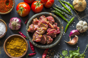 A plate of fresh tilapia mutton curry cut surrounded by ingredients garlic, tomatoes, dry red chillies, green chilly, turmeric for preparing mutton pepper dry fry recipe